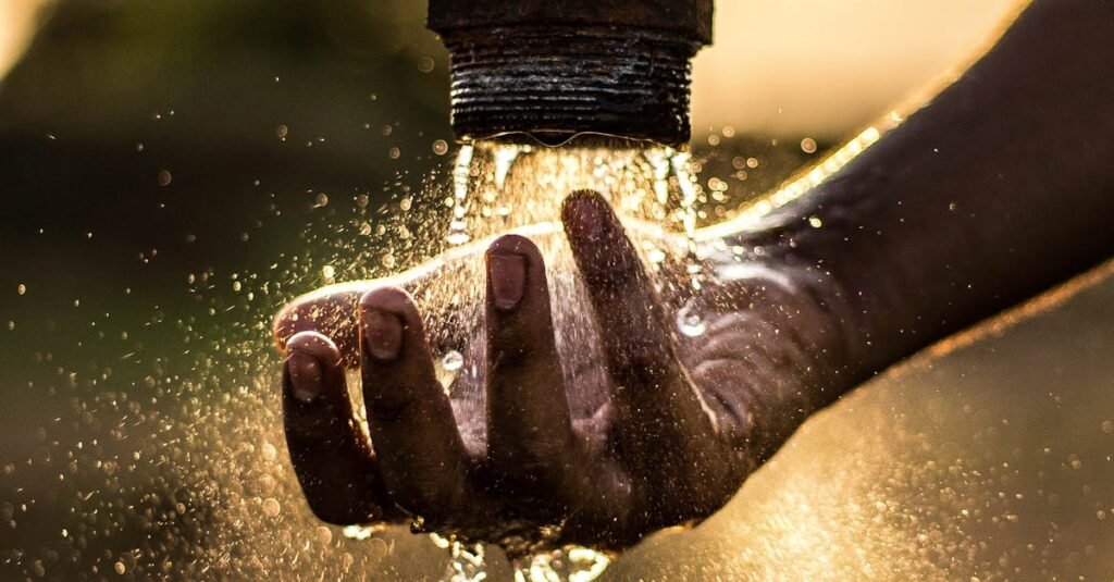 A person washes their hand under a rustic water pipe, capturing the essence of fresh water at sunset.