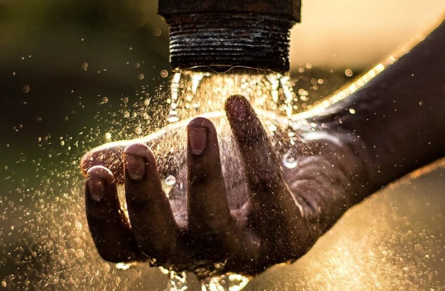 A person washes their hand under a rustic water pipe, capturing the essence of fresh water at sunset.